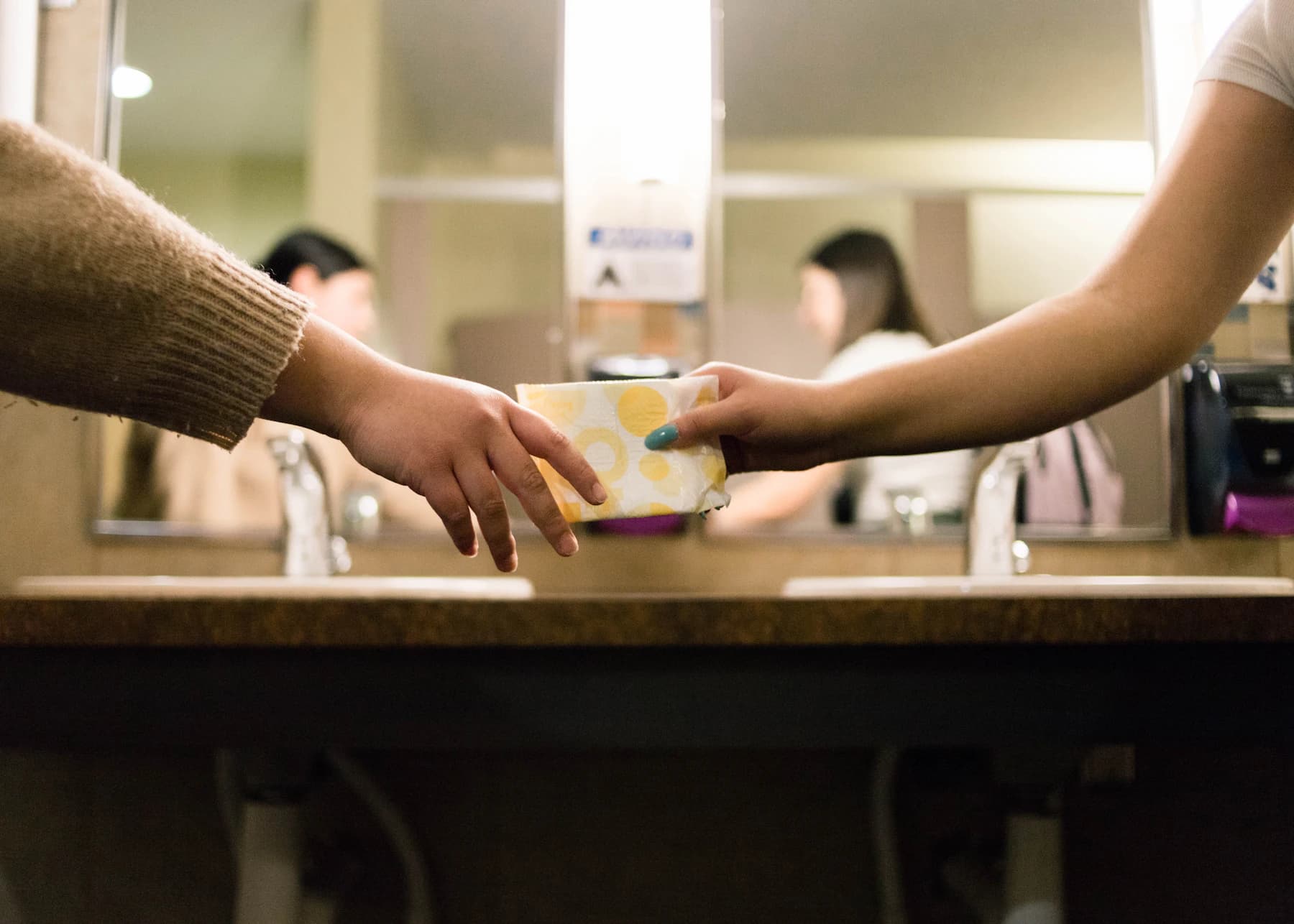 A period product being passed to someone in a bathroom